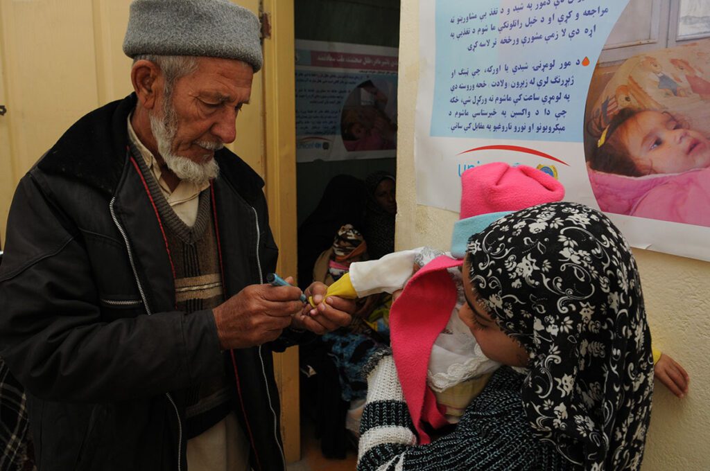 An older man wearing a cap and jacket marks the finger of a young child with a pen, while the child, wearing a knit cap and a patterned headscarf, opens her mouth to receive drops from a small vaccine vial. Other people are visible in the background near a doorway, and educational posters with text in a non-Latin script hang on the wall behind them.