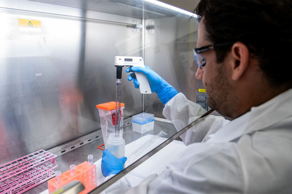 Wastewater Surveillance Detects New Flu Variant a Month Before It Shows Up in Hospitals A laboratory scientist wearing protective gloves and glasses works inside a biosafety cabinet, handling wastewater samples and analytical equipment. The scientist operates instrumentation and manipulates sample containers with sample racks visible on the work surface. Laboratory materials and equipment are organized within the contained workspace. Safety signage and protective measures are visible in the background.