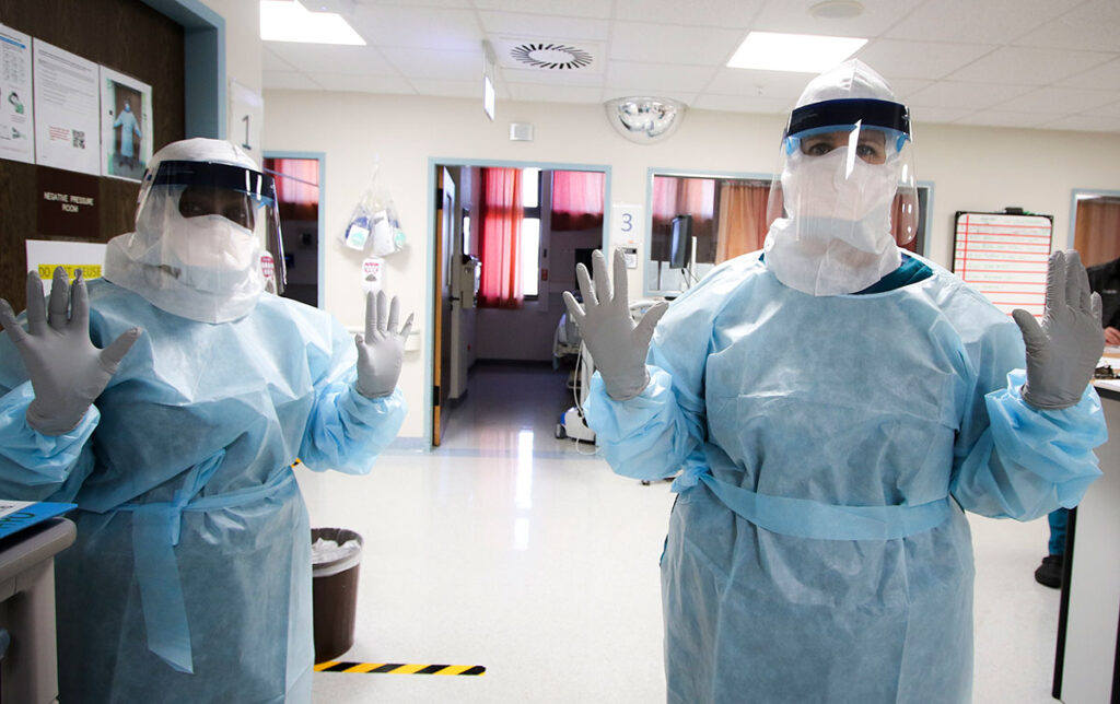 Two healthcare workers stand in an intensive care unit wearing full protective equipment including face shields, protective gowns, and gloves. Both are positioned facing the camera with hands raised, demonstrating the complete protective setup required for COVID-19 patient care. Medical equipment, monitoring stations, and hospital facility infrastructure are visible in the background. The scene depicts the level of protection necessary for frontline clinical work during the pandemic.