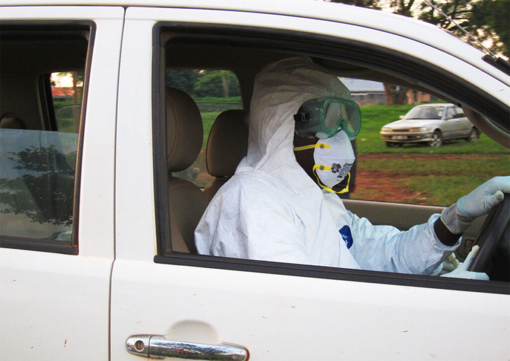 Congo’s Ebola Outbreak Could Be Three Times Its Confirmed Size as Vaccine Doses Finally Arrive An emergency responder wearing full protective equipment including a hooded suit, goggles, and respirator mask sits in the driver's seat of a vehicle, gripping the steering wheel with gloved hands. The vehicle is in motion, viewed through the open driver's side window. Rural roadside scenery including grass, trees, and another vehicle is visible in the background.