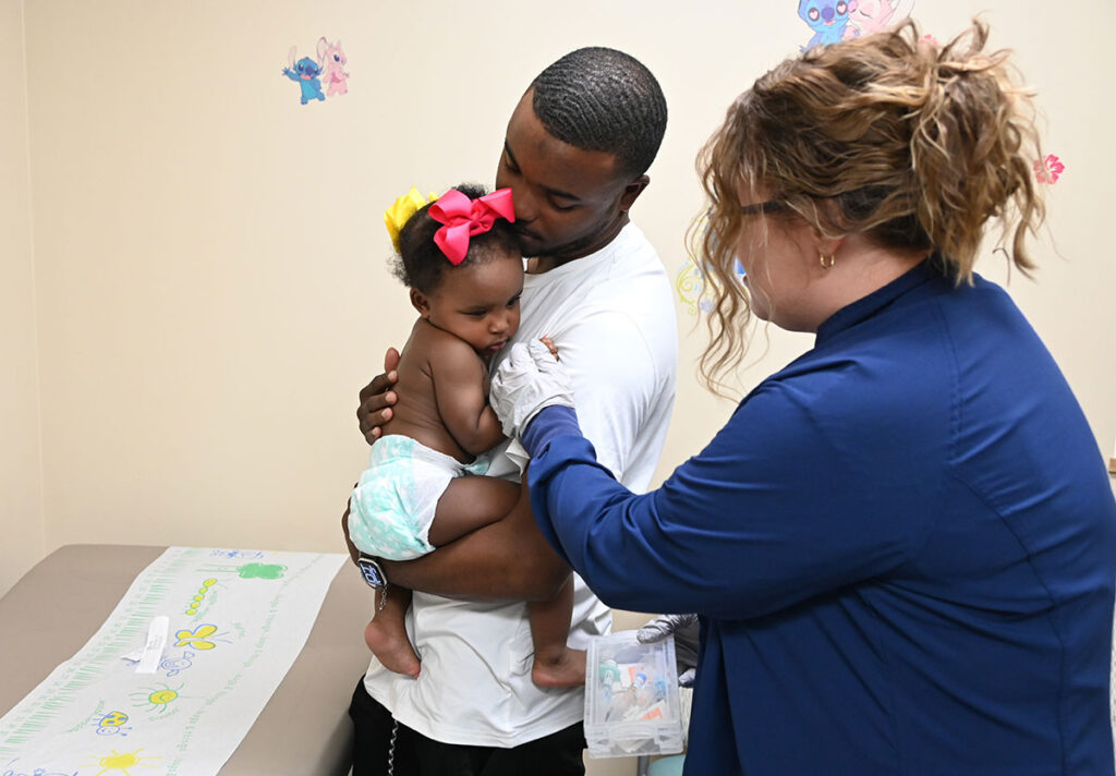 Executive Order on Vaccines Bypasses an Evidence-Based Process That Has Kept Kids Safe A nurse in a navy blue top administers an injection into the upper arm of an infant, who wears a bow in her hair and is held against the chest of a man in a white shirt. The man kisses the top of the infant's head while cradling her. A paper-covered exam table with decorative drawings sits nearby, and children's wall decals are visible on the wall behind them.