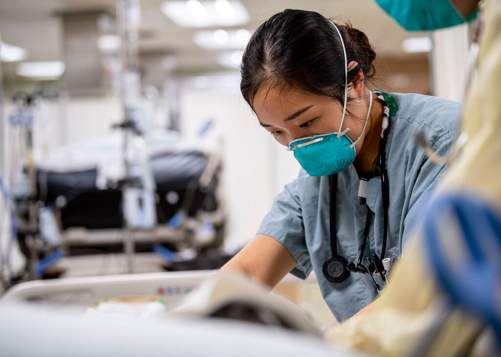 South Korea’s Bioterrorism Response Training Program Shows Strong Gains A healthcare worker wearing a teal face mask, blue scrubs, and a stethoscope around her neck leans over a hospital bed, attending to a patient. Her dark hair is pulled back and secured beneath the mask's straps. In the blurred background, medical equipment including IV poles and another hospital bed are visible in a ward setting.