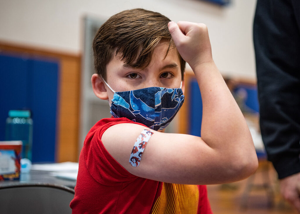 A young boy in a red shirt and a whale-print face mask turns to face the camera while flexing his bicep, showing a small bandage on his upper arm. He raises his other hand toward his forehead. In the blurred background, a water bottle and supplies sit on a table inside a gymnasium-style room with blue wall padding.