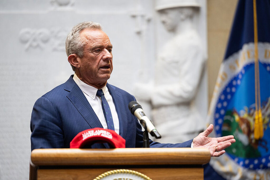 A man with gray hair speaks at a wooden podium, gesturing with one open hand while wearing a dark pinstriped suit, white shirt, and patterned tie. A red cap rests on the podium in front of him. Behind him, a carved stone relief and a state flag with a circular seal are visible, along with a microphone positioned near his face.