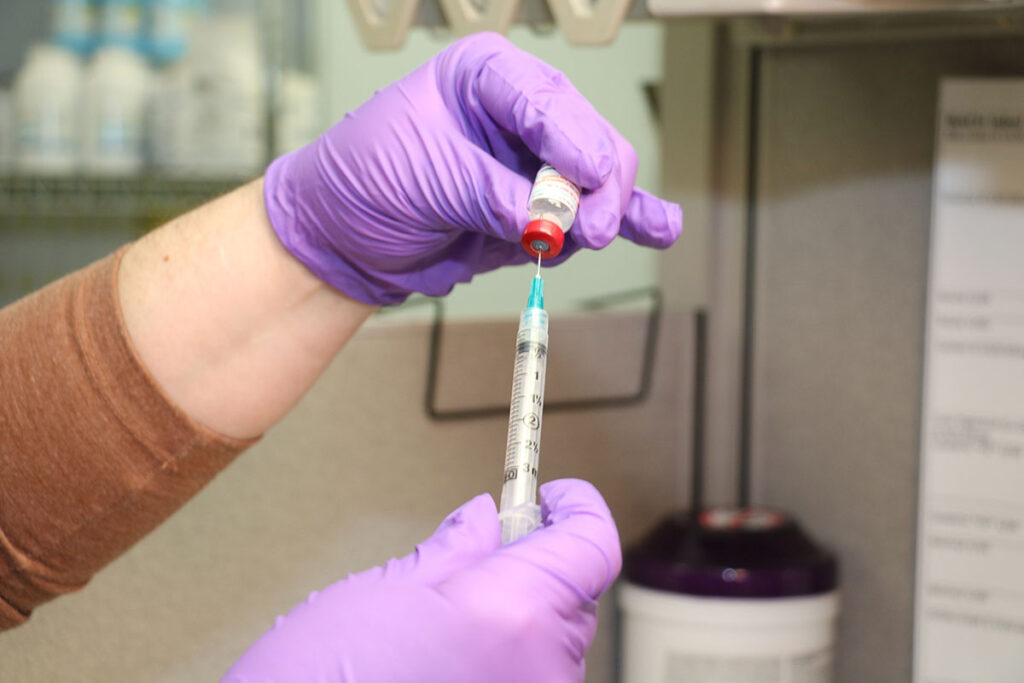 A close-up view of gloved hands wearing purple nitrile gloves, drawing liquid from a small vial into a syringe. One hand steadies the vial while the other holds the syringe with its plunger partially depressed, showing measurement markings. Shelves with supplies and a waste container are visible blurred in the background.