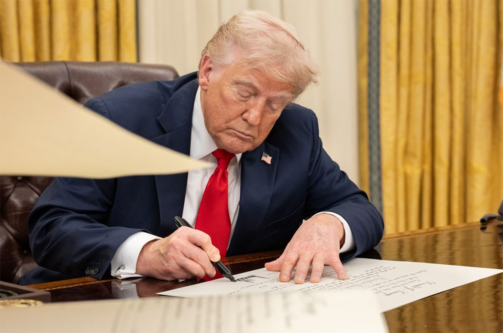 Trump’s Vaccine Executive Order Adds Chaos to an Already Strained System A man with light-colored hair, wearing a dark suit, white shirt, red tie, and a small flag pin, sits at a desk signing a document with a pen. His other hand rests flat on the paper. Gold curtains are visible in the background behind a leather chair.