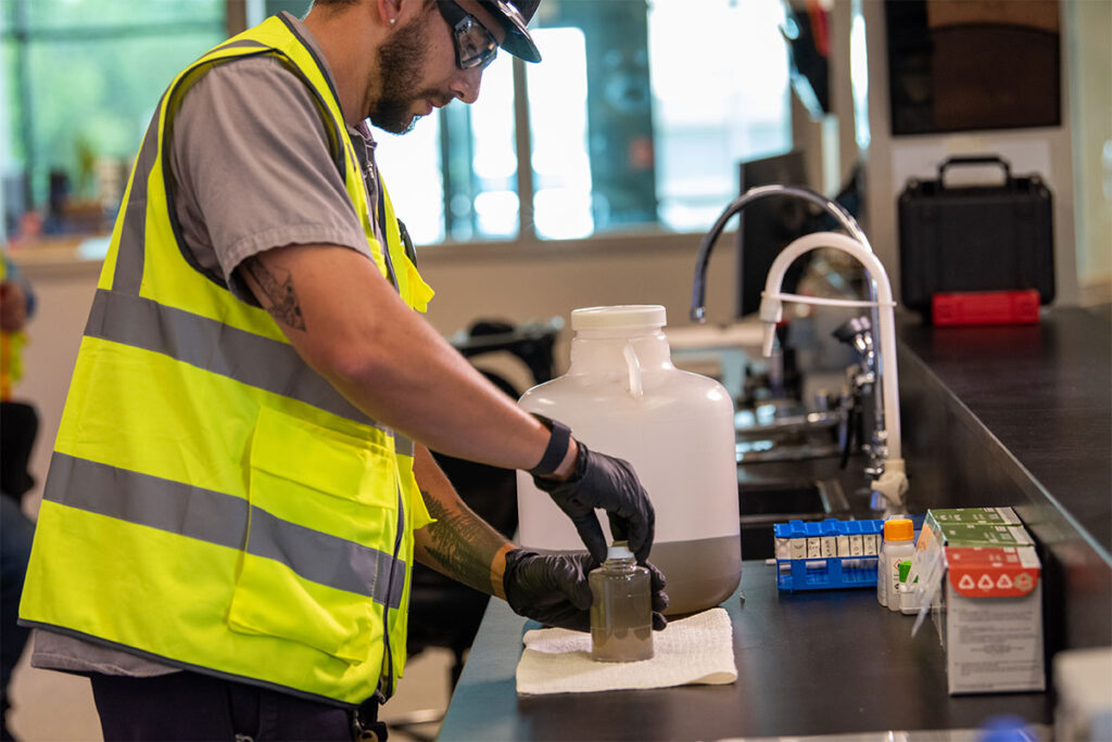 A man wearing a high-visibility safety vest, cap, and safety glasses stands at a lab counter, using gloved hands to handle a small sample bottle filled with murky liquid, positioned on a paper towel next to a large jug of similar liquid. A sink with a gooseneck faucet, a rack of test tubes, and various labeled testing supplies and boxes are arranged on the counter nearby.