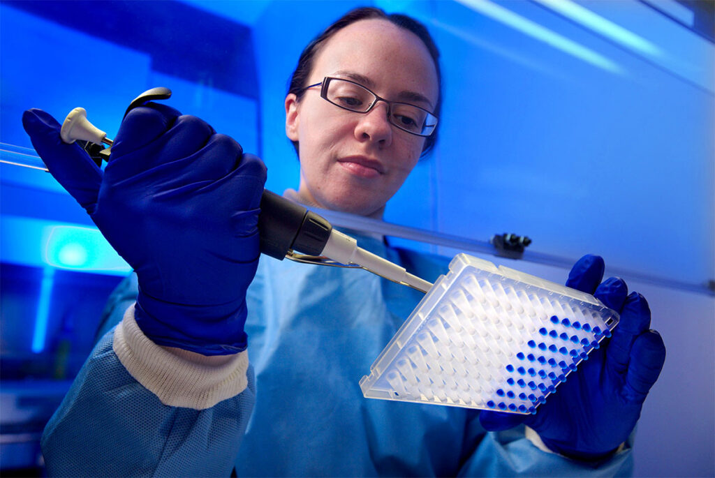 Newer Antibiotics Are Approved Without Testing Them on the Sickest Patients Who’ll Need Them A researcher wearing glasses and a blue protective gown and gloves holds a mechanized pipette in one hand while pipetting solution into a 96-well plate held in the other hand, with several wells already filled with a dark liquid. The scene is lit with a blue-toned glow inside a ventilated laboratory hood.