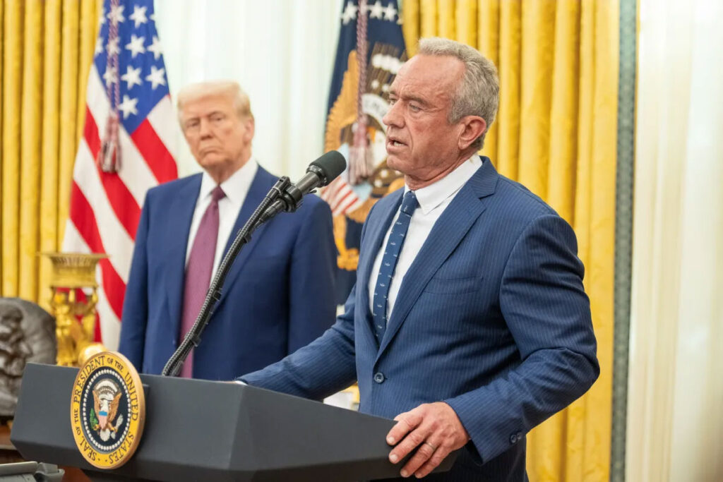 CDC vs. Pennsylvania: Why Conflicting Public Health Messages Jeopardize Public Safety A man with gray hair speaks into a microphone at a podium bearing the presidential seal, wearing a dark pinstriped suit and patterned tie. Behind him, another man in a dark suit and tie stands watching, positioned in front of an American flag and a presidential flag, with gold curtains filling the background.
