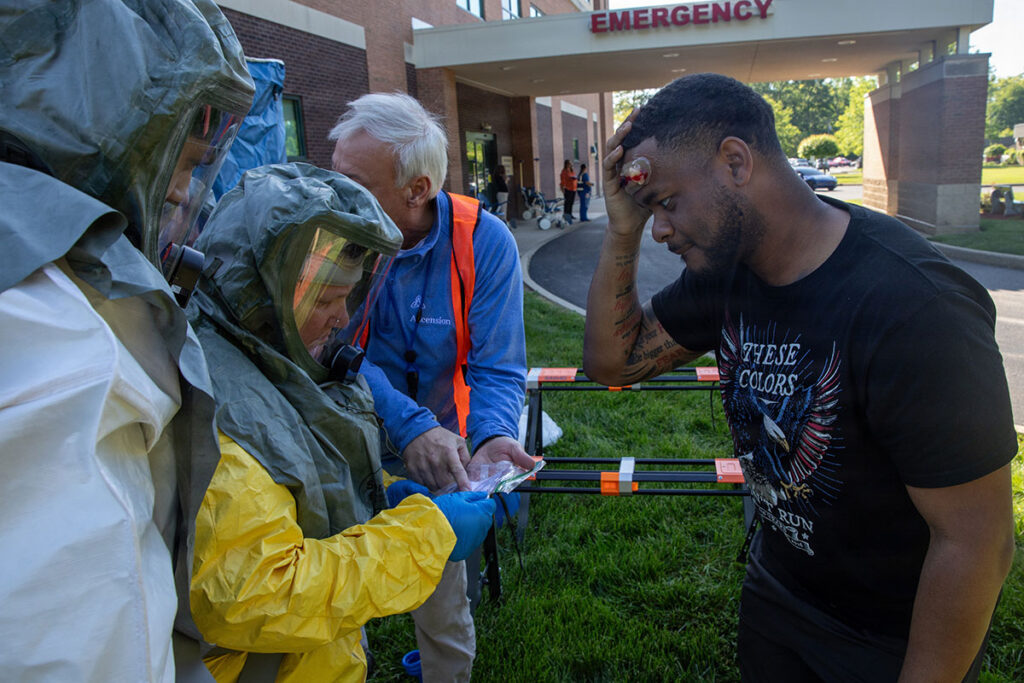 The Five Assumptions That Fail Hospitals During a Chemical Disaster Two people in olive-green hooded protective suits with face shields, one wearing bright yellow gloves, examine a small item held up between them, while a man in a blue jacket and orange safety vest looks on. To their right, a man in a black t-shirt stands with his hand pressed to his forehead, where a small bandage is visible, appearing to portray an injured patient. An orange-and-black barrier stands on the grass nearby, and a hospital building with an "EMERGENCY" sign is visible in the background.