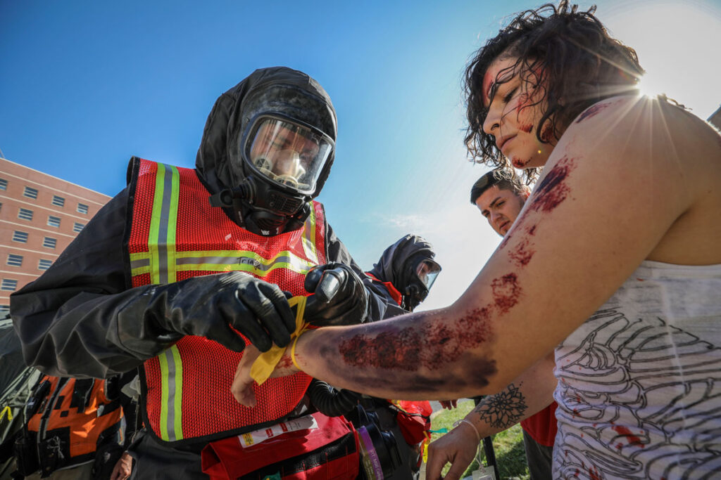 89 Million Americans Live Near High-Risk Chemical Facilities With No Federal Way to Vet Staff for Terrorist Ties A person in a hazmat suit with a full face respirator and a red high-visibility vest ties a yellow ribbon around the wrist of a woman whose arm and shoulder are marked with simulated wounds and dried blood, portraying an injury victim. Another responder in protective gear stands behind them. A brick building and clear sky form the background.