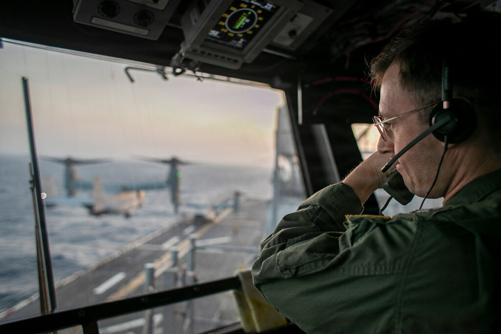 Pentagon Pain-Medication EUA Pathway Opens Amid Ongoing Iran Conflict A man wearing a green flight suit, glasses, and a headset with an attached microphone looks out through an open cockpit-style window toward the deck of a ship, one hand raised to his headset. Through the window, a tiltrotor aircraft with its propellers angled upward sits on the flight deck, with the ocean stretching to the horizon under a soft, hazy sky.