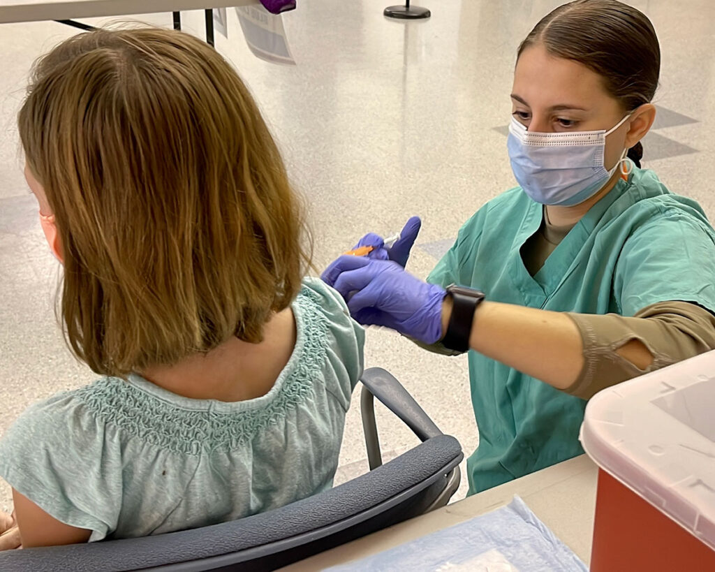 Debunking the Myths That Are Eroding America’s Vaccine Defenses A healthcare worker in green scrubs, a surgical mask, and purple gloves administers an injection into the upper arm of a seated child with shoulder-length hair, seen from behind. A red sharps container sits on the counter in the foreground, and additional medical supplies are visible on a table in the blurred background.
